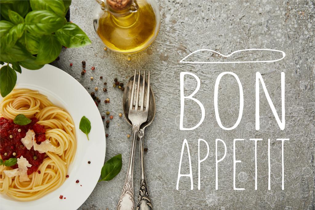 bovenaanzicht van heerlijke spaghetti met tomatensaus op bord bij basilicumbladeren, olie en bestek op grijs getextureerd oppervlak met bon appetit illustratie - Foto, afbeelding