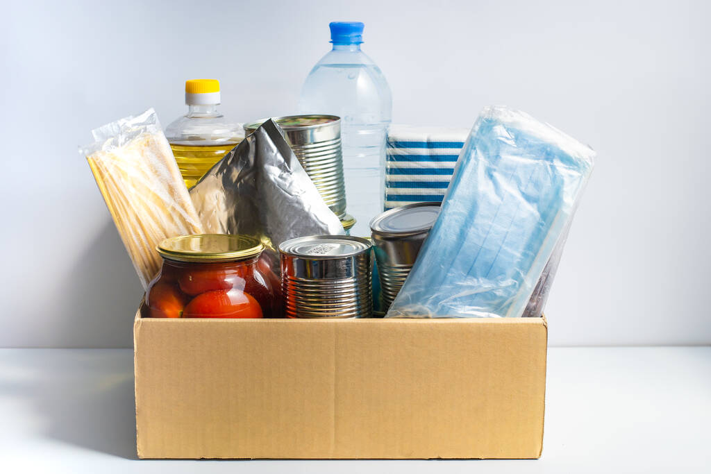 Box with food for a charity donation. Canned food, pasta, pickled cucumbers, vegetable oil, tea. Paper napkins, protective masks. Food delivery or food donation concept. Coronavirus quarantine. - Photo, Image