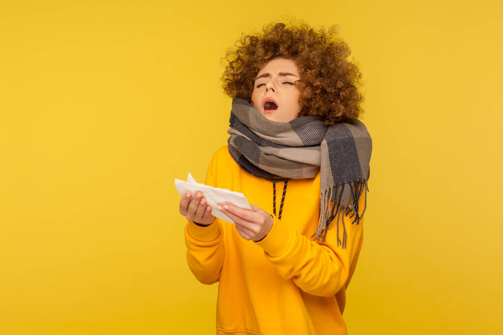Seasonal influenza. Flu-sick woman with fluffy curly hair, wrapped in warm scarf, open mouth to sneeze in napkin, feeling unwell, coughing and blowing nose. indoor studio shot, yellow background - Photo, Image