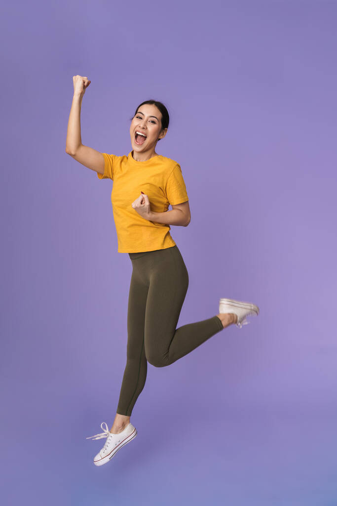 Volle Länge einer fröhlichen jungen hübschen Sportlerin, die isoliert über violetten Hintergrund springt und mit Hanteln trainiert - Foto, Bild