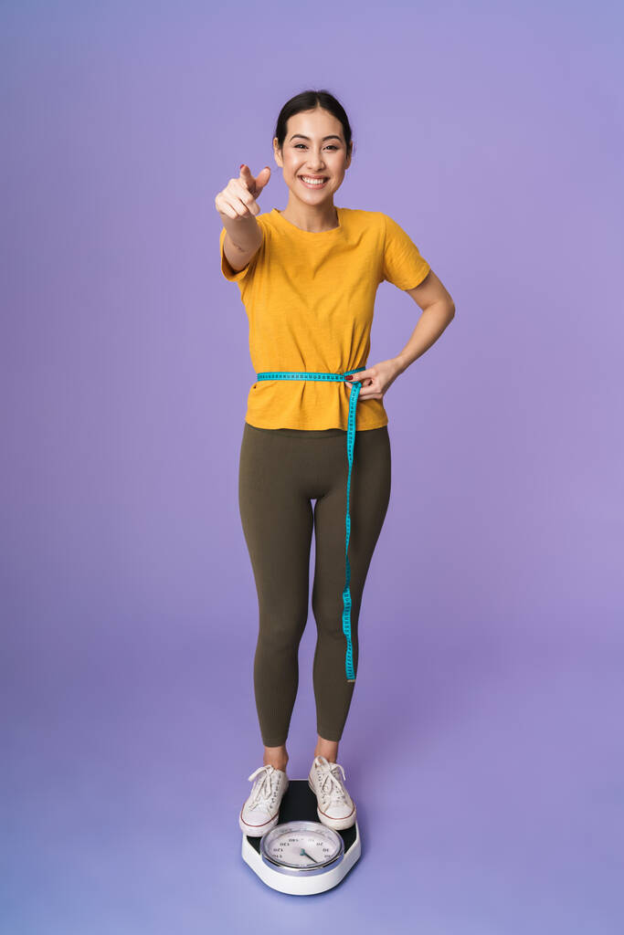 Volledige lengte van een vrolijke jonge mooie sportvrouw op schalen geïsoleerd over violette achtergrond, met meetlint rond haar middel, wijzende vinger naar camera - Foto, afbeelding