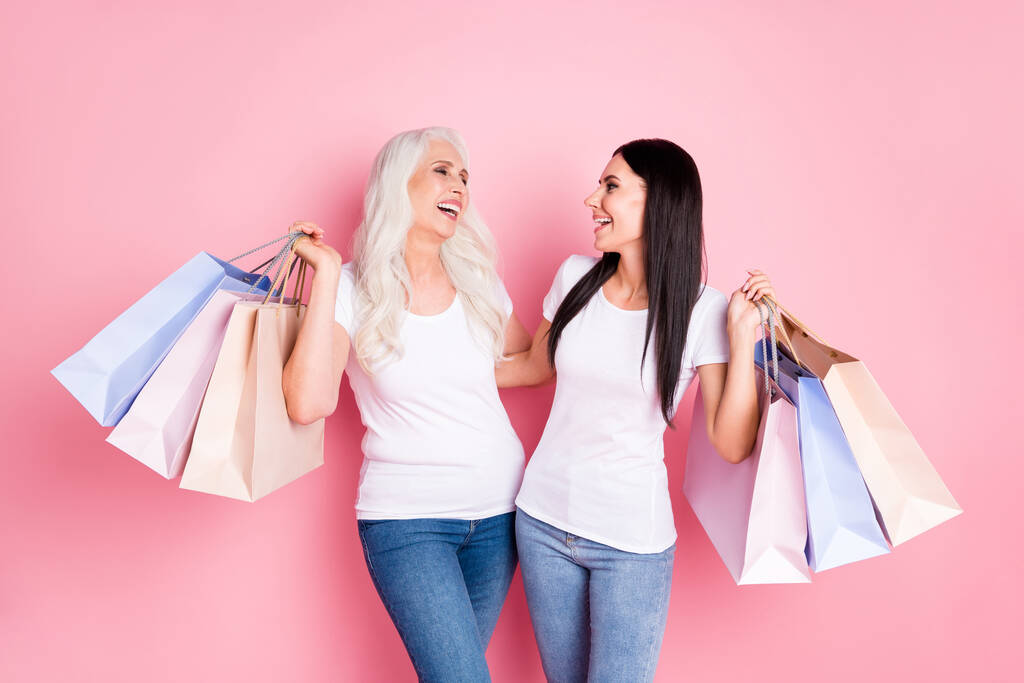 Kuva hauska ikäinen äiti nuori tytär hyvät pitää monia myymälää pakkauksissa käydä ostoksilla yhdessä riemuiten kulumista valkoinen t-paidat farkut eristetty pastelli vaaleanpunainen väri tausta
 - Valokuva, kuva