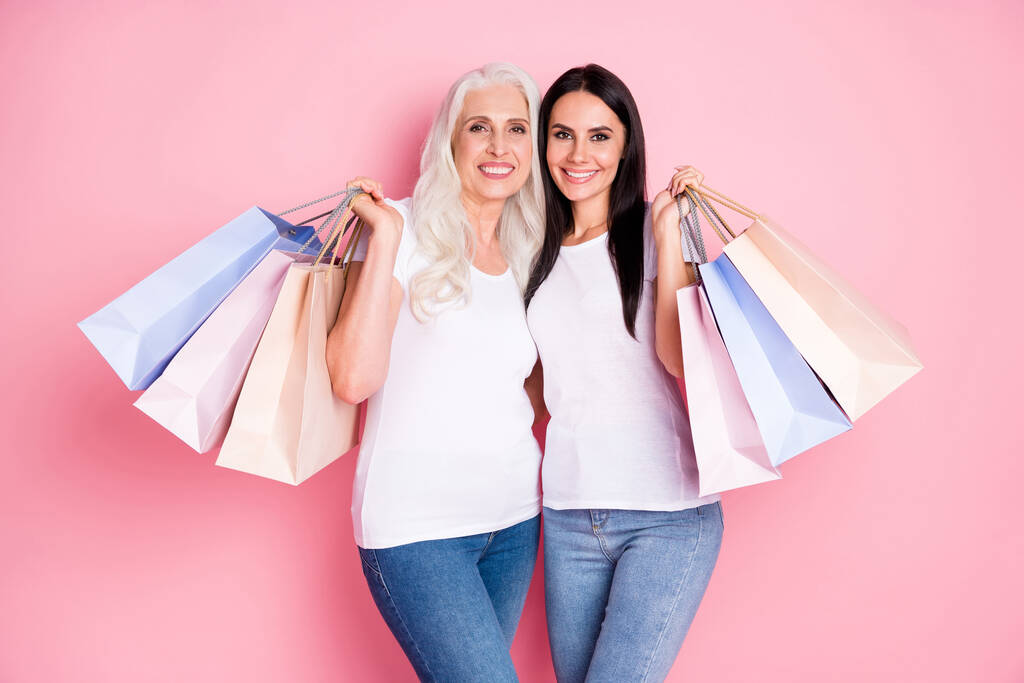 Foto de alegre anciana madre joven hija damas sosteniendo muchos paquetes de compras asisten a la tienda juntos usan camisetas blancas jeans aislados pastel rosa color fondo
 - Foto, imagen