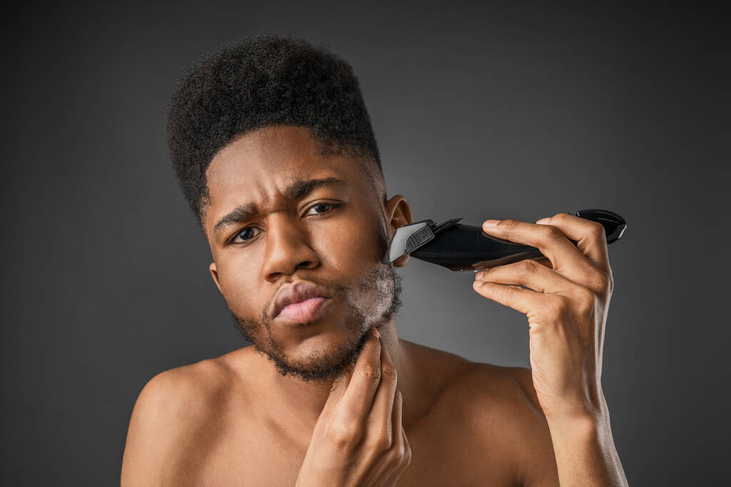 Крупним планом молодий африканський чоловік голиться борода з електричною бритвою на темно-сірому фоні
 - Фото, зображення