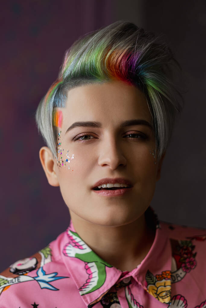 Portrait of a young beautiful girl in a pink shirt on a gray background with dyed hair. Short haircut pixie or bob. Rainbow coloring and sequins on the face. - Photo, Image