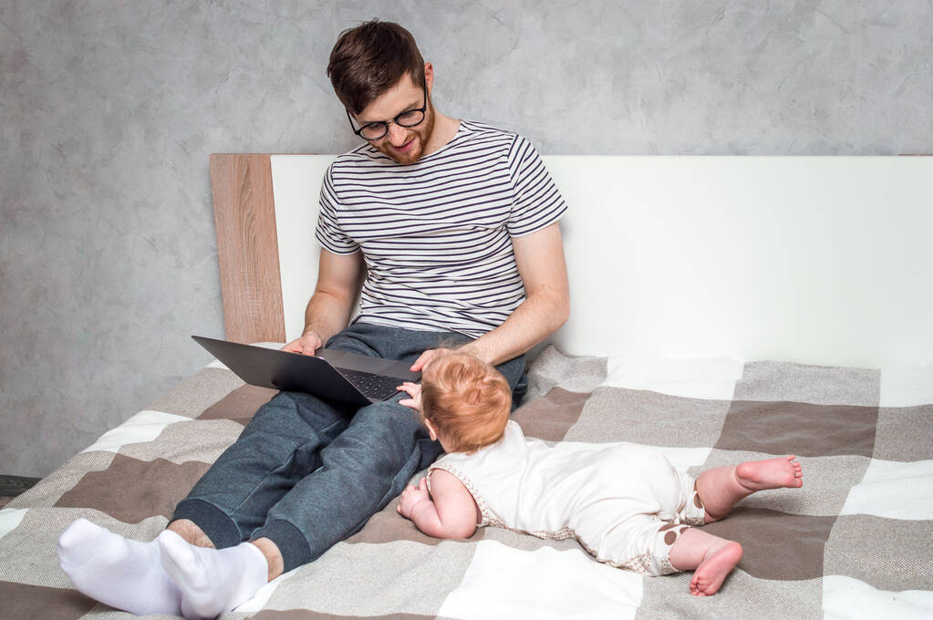 Een jongeman die thuis aan een laptop werkt en een baby ernaast. Concept Op afstand werken - Foto, afbeelding