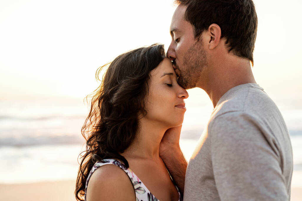 Milostivý mladý muž líbání své ženy na čelo, zatímco stojí spolu na pláži za soumraku - Fotografie, Obrázek