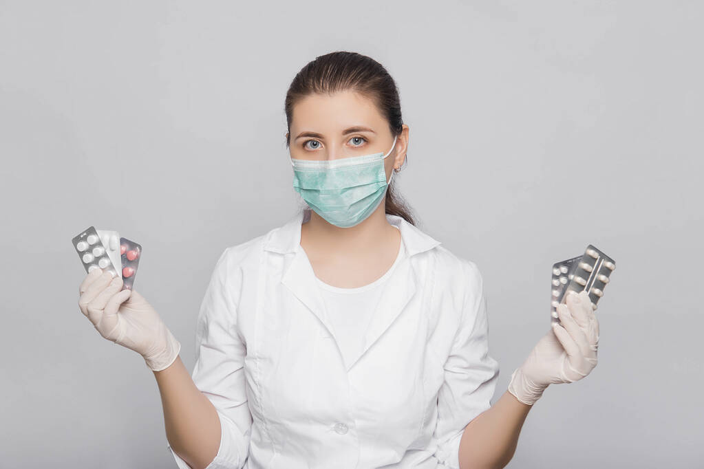 Молода жінка-лікар в білому пальто, рукавички і маска тримає таблетки в руках, стоячи на білому тлі
. - Фото, зображення