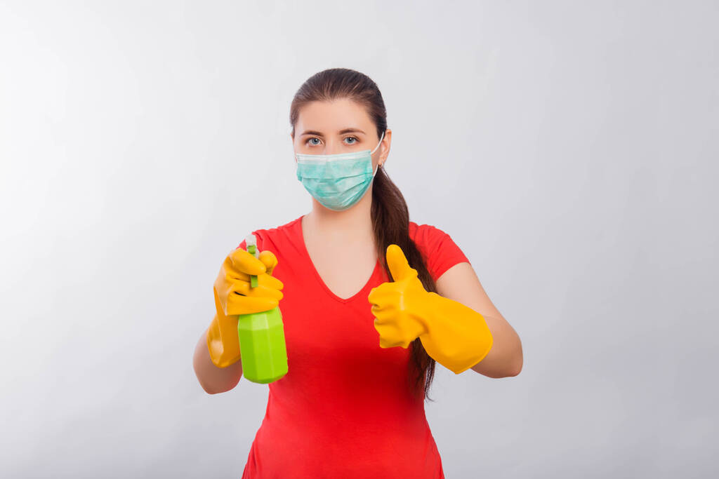 Uma jovem mulher com uma máscara e luvas amarelas segura uma garrafa de spray verde em suas mãos, isolada em um fundo branco. Arte conceitual para salas de limpeza e prevenção de doenças virais
. - Foto, Imagem