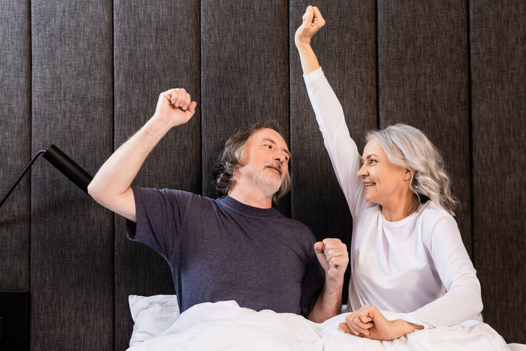 счастливые зрелые мужчина и женщина с руками над головой смотрят друг на друга в спальне
 - Фото, изображение