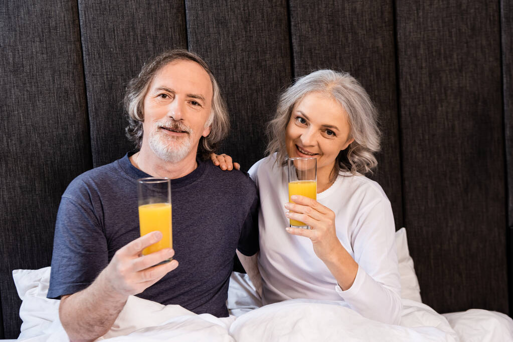 šťastný dospělý pár drží brýle s pomerančovým džusem v posteli - Fotografie, Obrázek