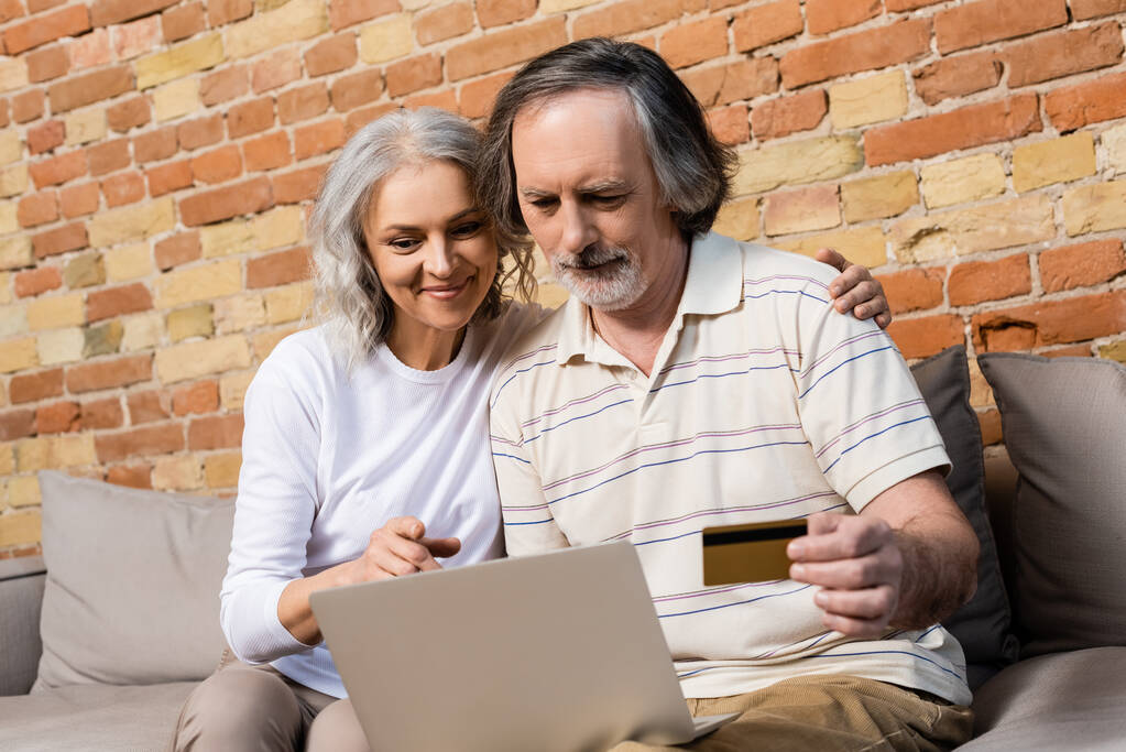 вибірковий фокус зрілої жінки, дивлячись на ноутбук біля чоловіка середнього віку з кредитною карткою
  - Фото, зображення