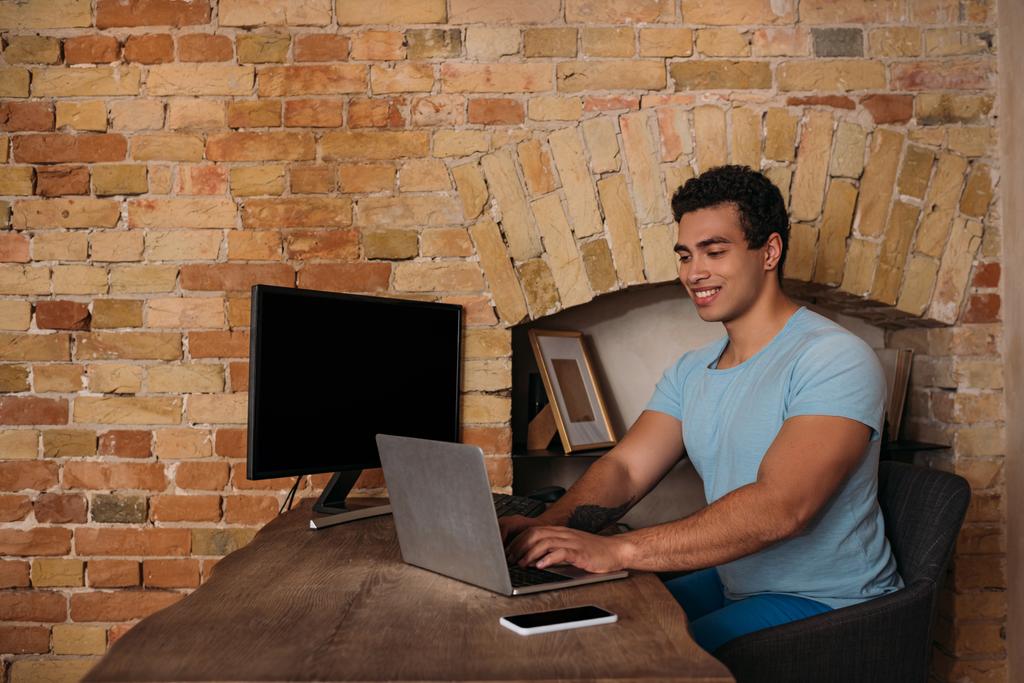 sonriente freelancer de carrera mixta que trabaja con computadora portátil, computadora y teléfono inteligente en la oficina en el hogar durante el autoaislamiento
 - Foto, imagen