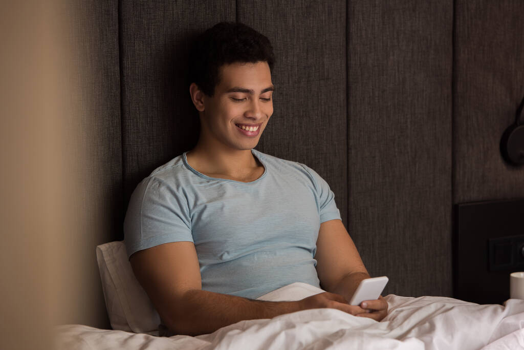 красивый улыбающийся смешанной расы человек, использующий смартфон в постели на карантине
 - Фото, изображение