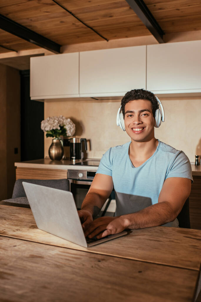 sonriente hombre de raza mixta escuchar música con auriculares y el uso de ordenador portátil durante el autoaislamiento en la cocina
 - Foto, Imagen