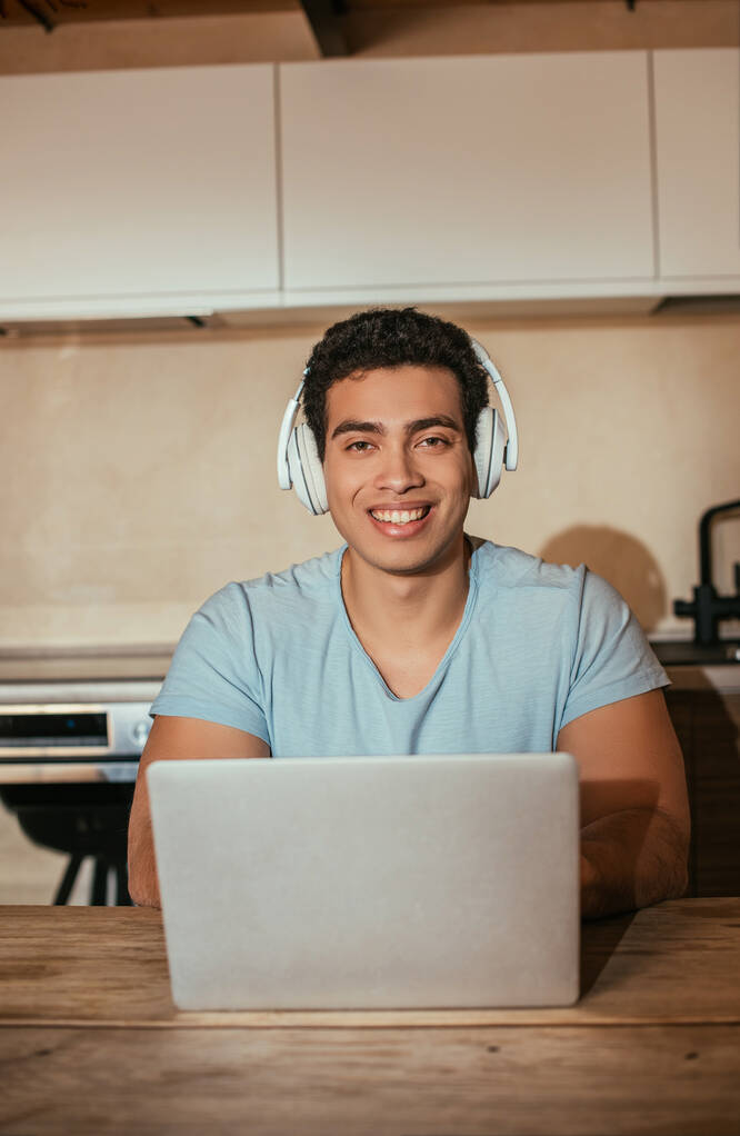 hymyilevä bi-rotu mies kuuntelee musiikkia kuulokkeilla ja kannettavan tietokoneen avulla itsensä eristämisen aikana
 - Valokuva, kuva