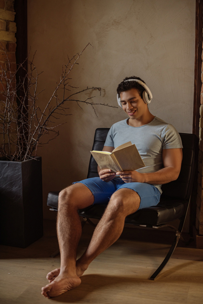 happy mixed race man reading book while listening music with headphones at home during quarantine - Photo, Image