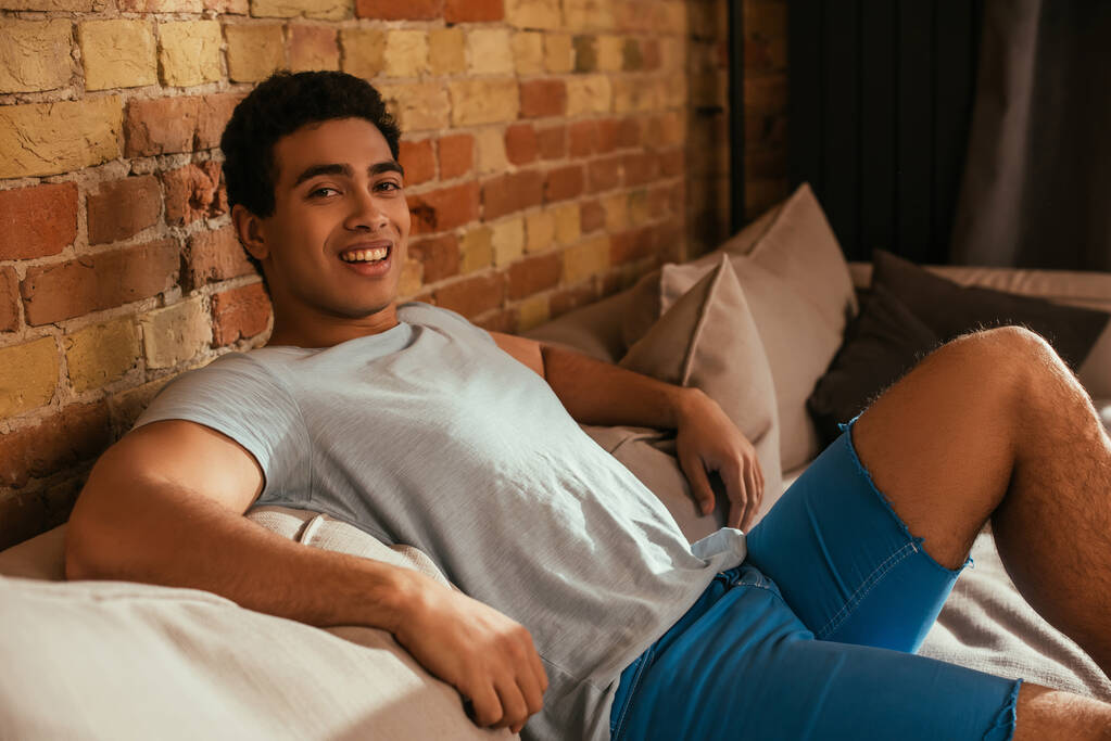 uomo di razza mista sorridente agghiacciante sul divano in soggiorno durante la quarantena
 - Foto, immagini