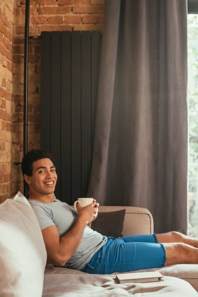 uomo di razza mista sorridente che tiene una tazza di caffè sul divano con libro durante l'autoisolamento
 - Foto, immagini