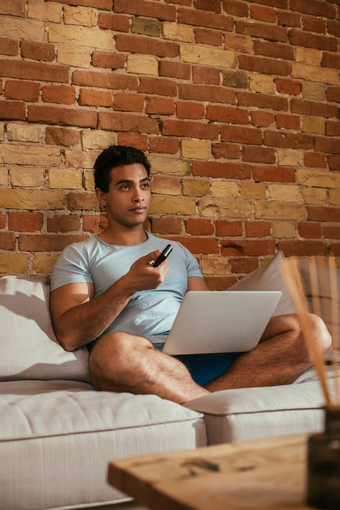 Dizüstü bilgisayarlı melez adam uzaktan kumandayı tutuyor ve kendini izole ederken oturma odasında televizyon izliyor. - Fotoğraf, Görsel