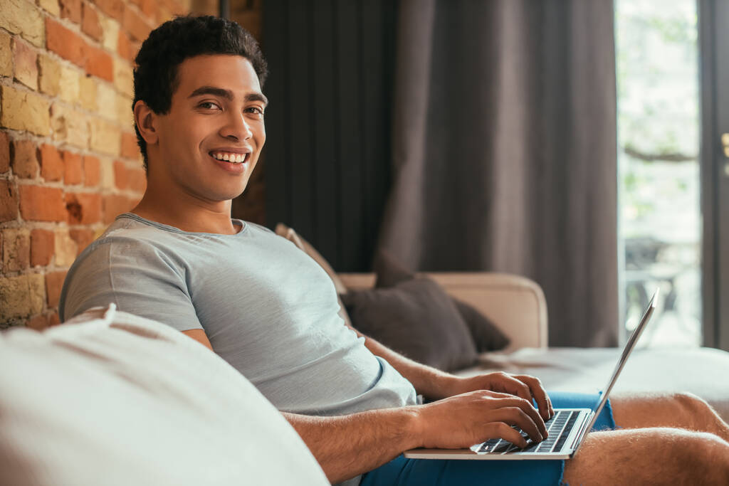 笑顔の混合人種の男は隔離されたソファの上でノートパソコンで凍えて - 写真・画像