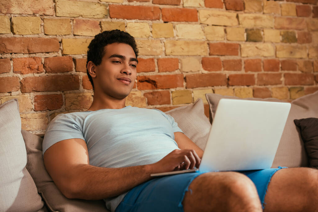 młody bi-rasowy mężczyzna chilling z laptopem na kanapie podczas samodzielnej izolacji - Zdjęcie, obraz