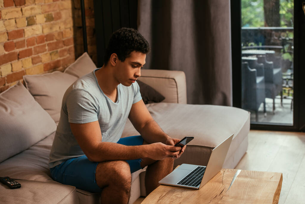 apuesto freelancer de raza mixta trabajando con smartphone y portátil en el sofá durante el autoaislamiento
 - Foto, Imagen