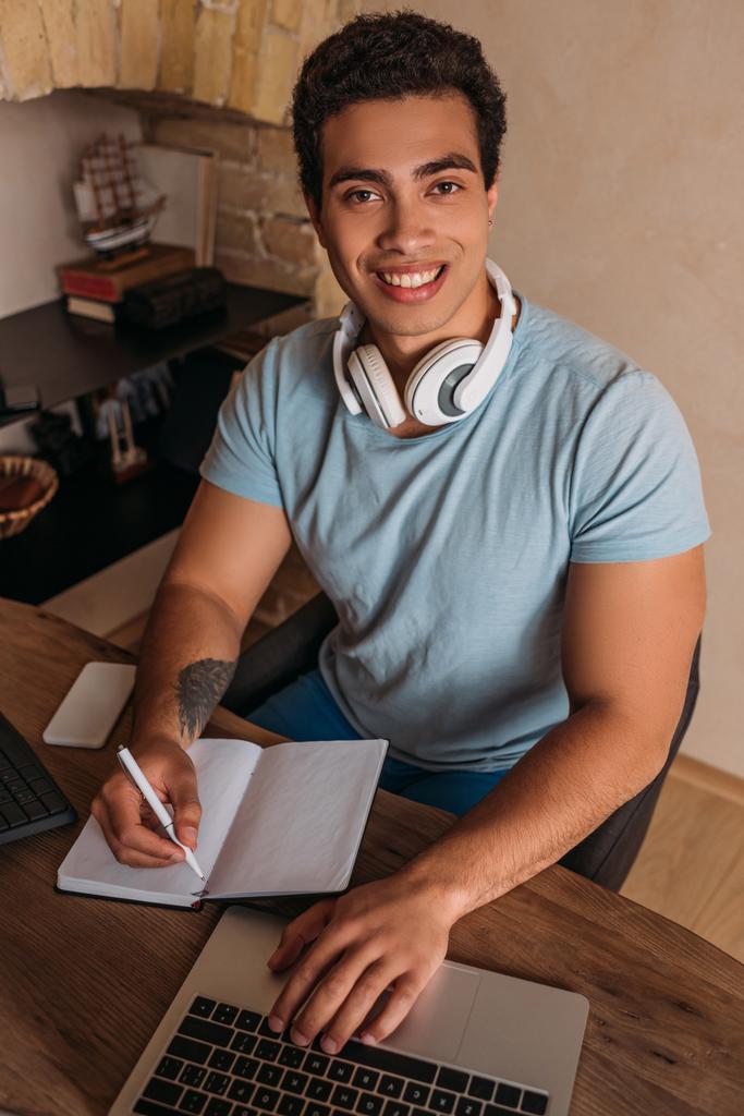 χαμογελαστός ελεύθερος επαγγελματίας μικτής φυλής που εργάζεται με φορητό υπολογιστή και σημειωματάριο στο γραφείο στο σπίτι κατά τη διάρκεια καραντίνας - Φωτογραφία, εικόνα
