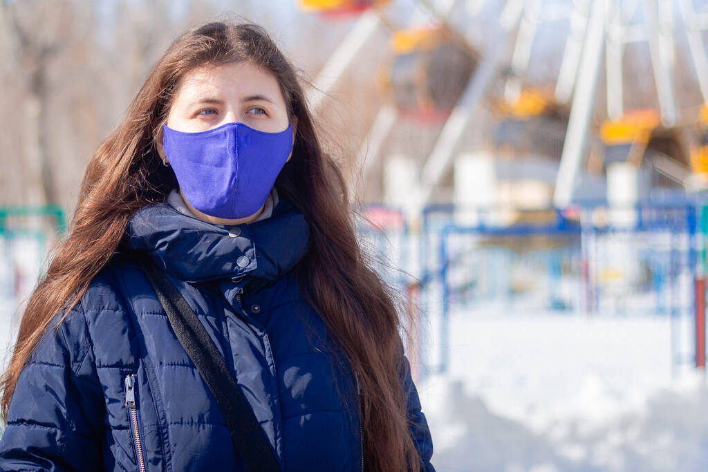 Portrait d'une fille vêtue d'une veste d'hiver avec une capuche et un masque de protection bleu sur son visage se promenant dans un parc d'attractions désert
. - Photo, image