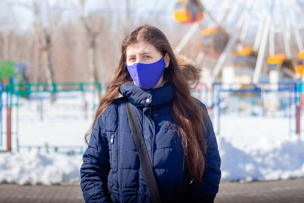 Πορτραίτο ενός κοριτσιού με χειμωνιάτικο μπουφάν με κουκούλα και μπλε προστατευτική μάσκα στο πρόσωπό της να περπατά σε ένα έρημο πάρκο διασκέδασης. - Φωτογραφία, εικόνα