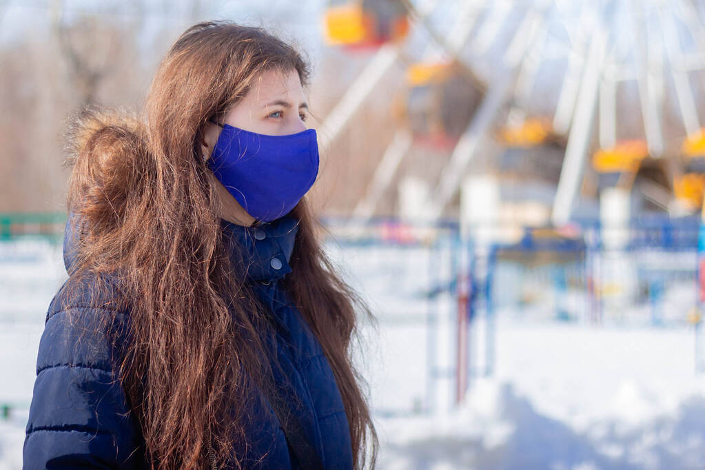 Porträt eines Mädchens in Winterjacke mit Kapuze und blauer Schutzmaske im Gesicht, das durch einen verlassenen Vergnügungspark spaziert. - Foto, Bild