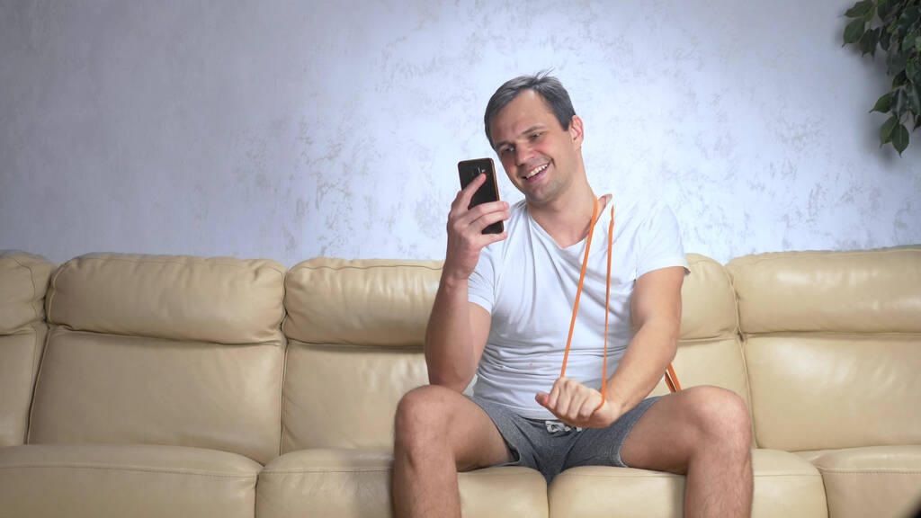 hombre en el sofá hace deportes con una banda elástica y utiliza un teléfono inteligente
 - Foto, imagen