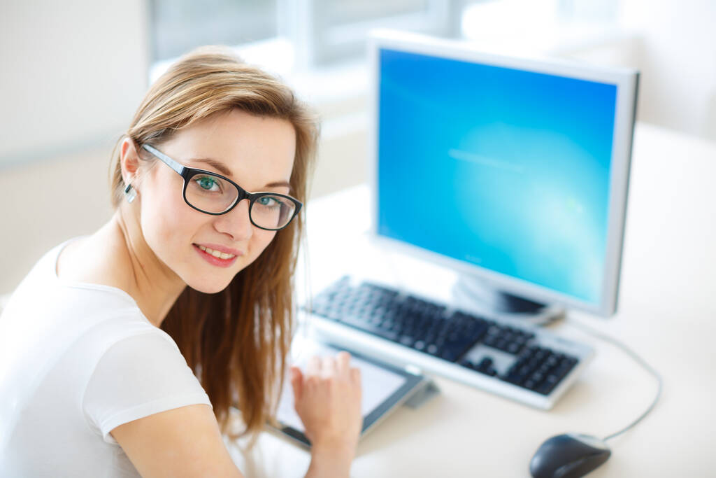 Χαμογελώντας φοιτήτρια / επιχειρηματίας χρησιμοποιώντας τον υπολογιστή tablet της και έναν επιτραπέζιο υπολογιστή, μένοντας ενημερωμένη, εργαζόμενη, κοιτάζοντας την κάμερα. - Φωτογραφία, εικόνα