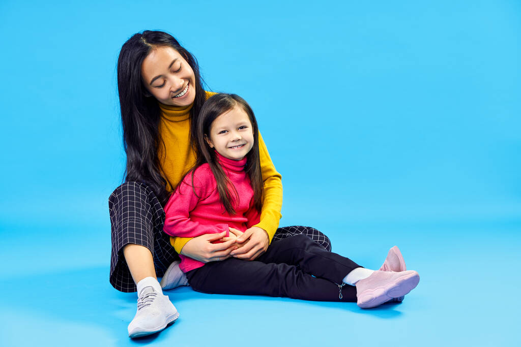 gelukkig glimlachen moeder knuffels haar kleine schattig dochter poseren op blauwe achtergrond - Foto, afbeelding