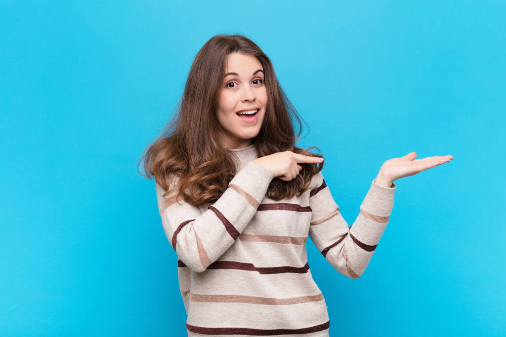 молодая красивая женщина весело улыбается и указывает на скопировать пространство на ладони на стороне, показывая или рекламируя объект на синей стене - Фото, изображение
