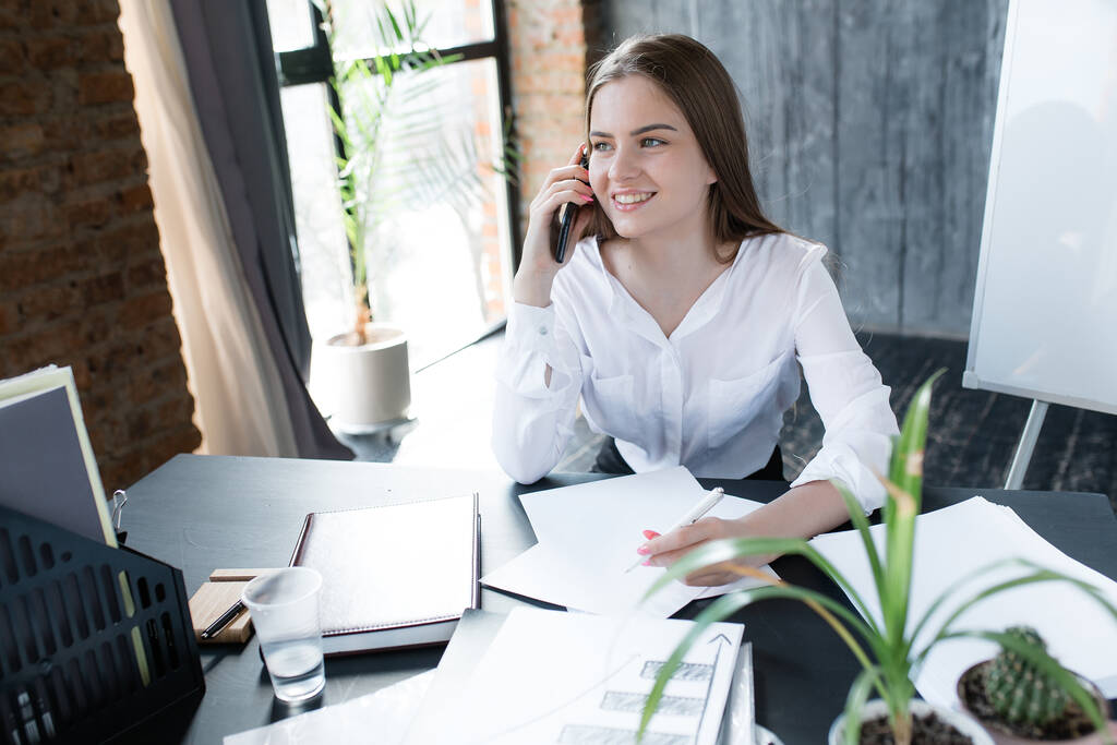 Chica europea hablando en un teléfono móvil y sonriendo. Trabajo en una oficina moderna con plantas, pizarra blanca con ventana panorámica
. - Foto, imagen