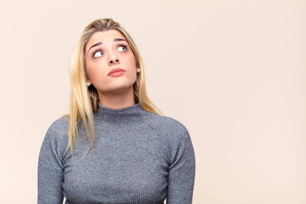 young pretty blonde woman with a worried, confused, clueless expression, looking up to copy space, doubting against flat wall - Photo, Image