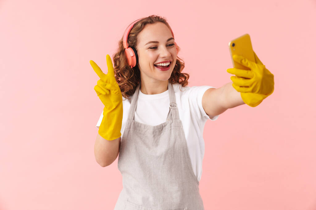 Photo d'une jeune femme au foyer heureuse et positive dans des gants isolés sur fond de mur rose prendre selfie par téléphone mobile écouter de la musique avec des écouteurs montrant la paix
. - Photo, image
