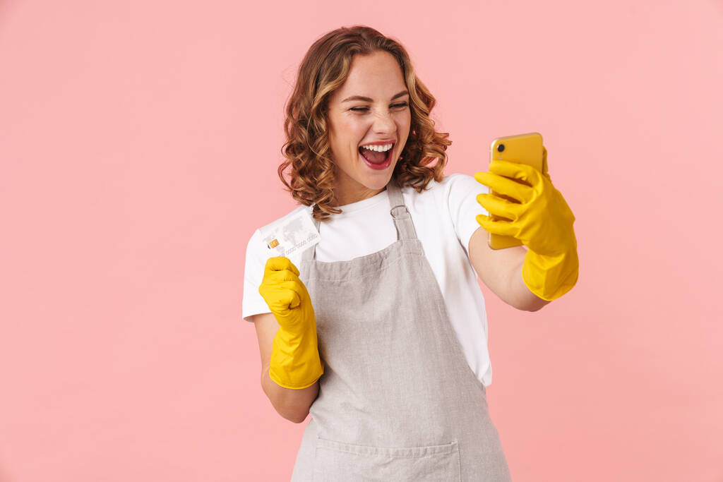Imagen de una joven ama de casa emocionada feliz con guantes que sostienen el teléfono móvil y la tarjeta de crédito aislados sobre fondo de pared rosa
. - Foto, Imagen