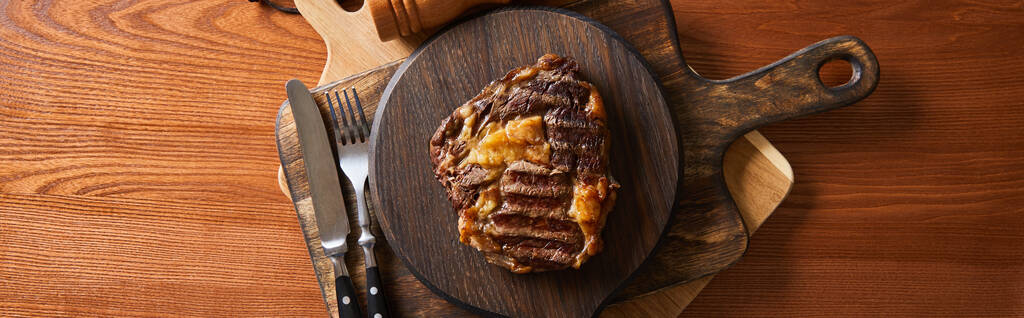 vrchní pohled na chutný grilovaný steak podávaný na dřevěné desce v blízkosti příborů, panoramatický záběr - Fotografie, Obrázek