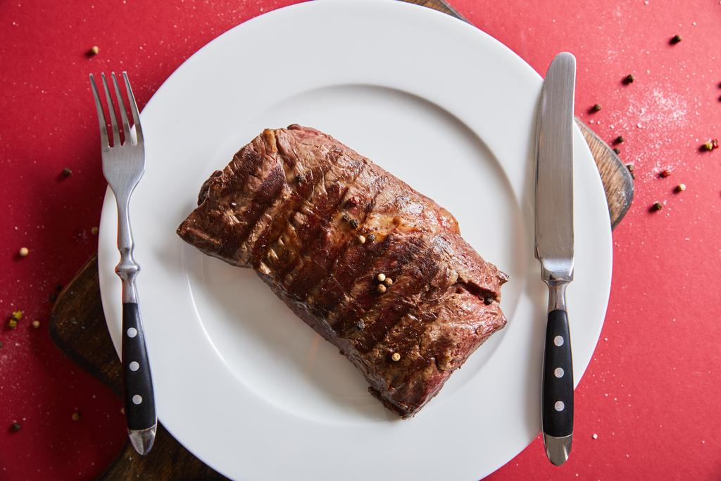 Blick von oben auf leckeres gegrilltes Steak serviert auf Teller auf Holzbrett auf rotem Hintergrund mit Pfeffer und Salz - Foto, Bild