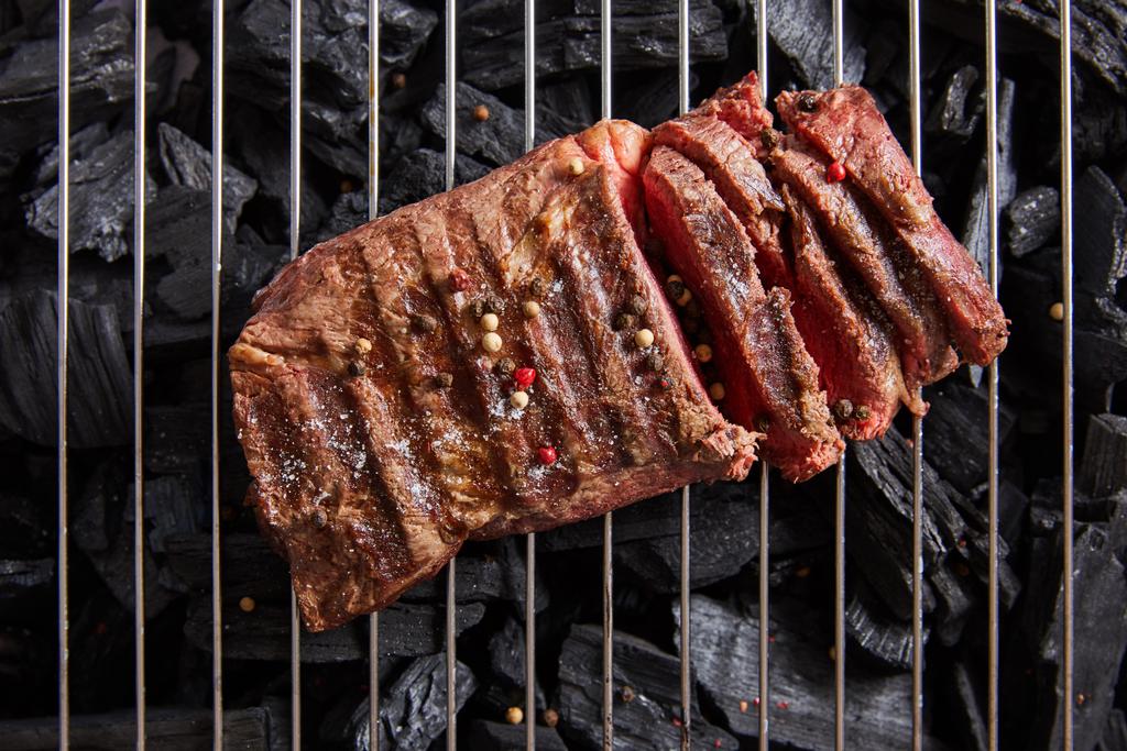 bovenaanzicht van gesneden verse gegrilde smakelijke steak met zeldzame roosteren en specerijen op rooster boven zwarte kolen - Foto, afbeelding