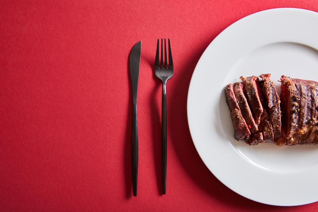Blick von oben auf leckeres Steak vom Grill serviert auf Teller mit Besteck auf rotem Hintergrund - Foto, Bild