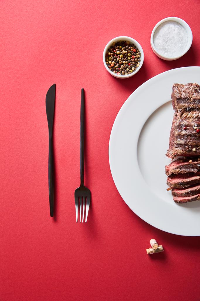 Blick von oben auf leckeres Steak vom Grill serviert auf Teller mit Besteck und Salz und Pfeffer in Schüsseln auf rotem Hintergrund - Foto, Bild