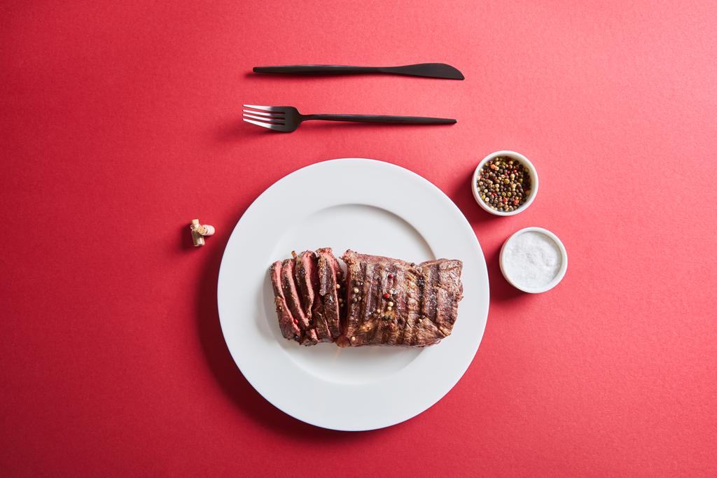 Blick von oben auf leckeres Steak vom Grill serviert auf Teller mit Besteck und Salz und Pfeffer in Schüsseln auf rotem Hintergrund - Foto, Bild