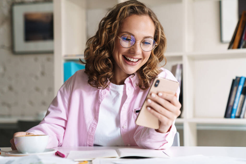 Фото улыбающейся милой женщины в очках, использующей мобильный телефон и пьющей кофе во время домашней работы в номере
 - Фото, изображение