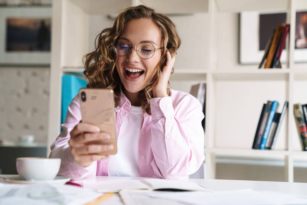 Fénykép mosolygós szép nő visel szemüveg, hogy videohívás a mobilon, miközben a házi feladatot a szobában - Fotó, kép