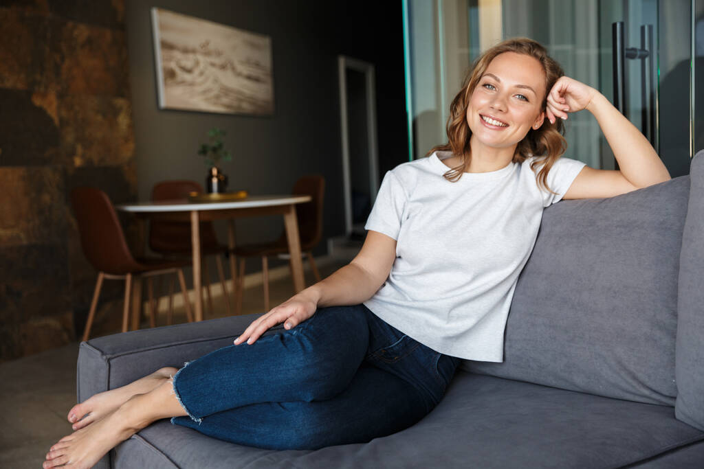 リビングでソファに腰かけながらカメラで微笑む基本的なTシャツ姿の幸せな白人女性の姿 - 写真・画像