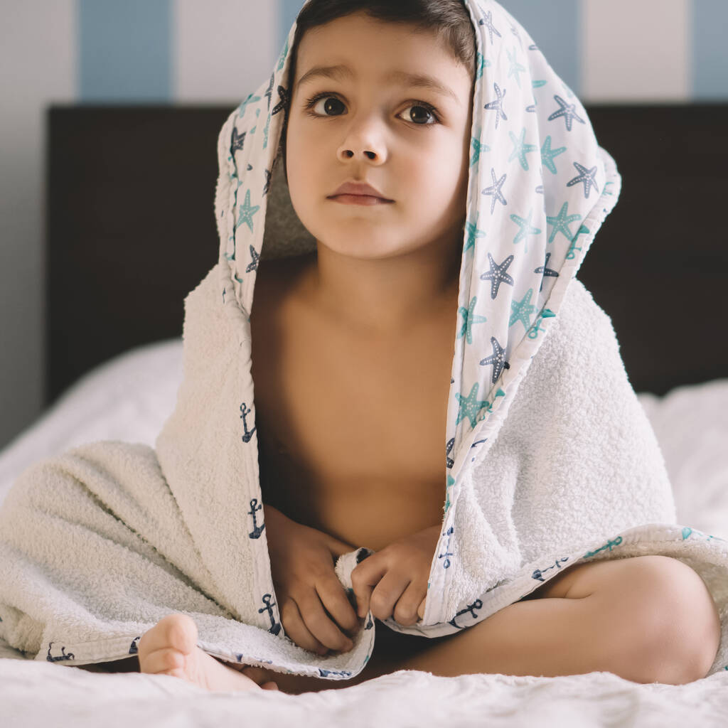 Αξιολάτρευτο αγόρι, καλυμμένο με κουκούλα πετσέτα, να κάθεται στο κρεβάτι και να κοιτάει αλλού - Φωτογραφία, εικόνα