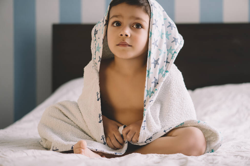 милый ребенок, покрытый полотенцем с капюшоном, сидит на кровати и смотрит в сторону
 - Фото, изображение